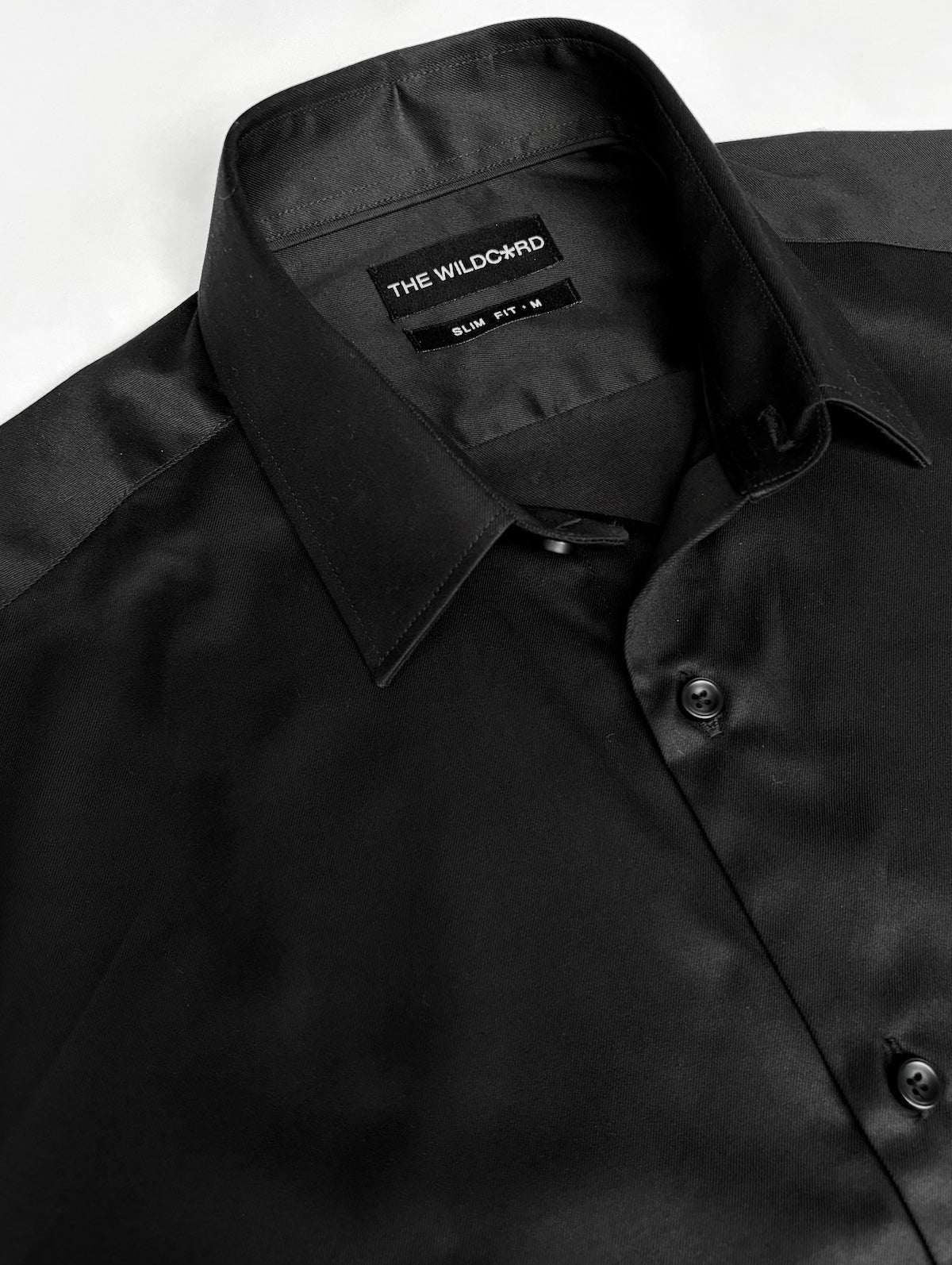 A close-up of a black long-sleeve smart shirt with a collar, showing the brand label 'THE WILDCARD' and fit and size label inside the collar.