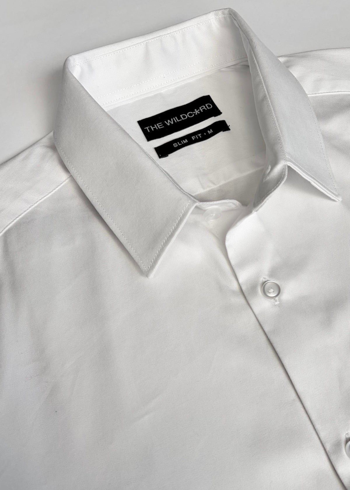 A close-up of a white long-sleeve smart shirt with a collar, showing the brand label 'THE WILDCARD' and fit and size label inside the collar.
