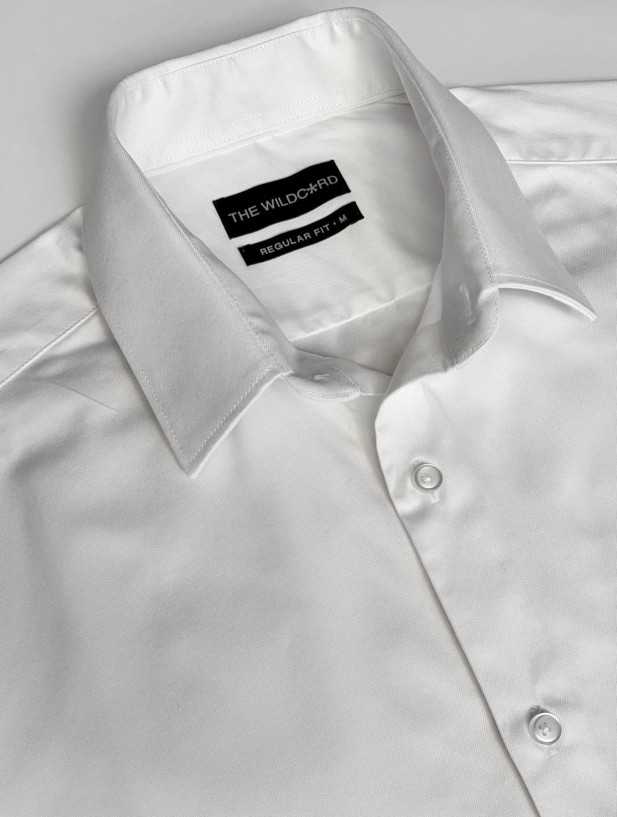 A close-up of a white long-sleeve smart shirt with a collar, showing the brand label 'THE WILDCARD' and fit and size label inside the collar.